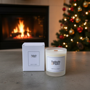 A Tadpole & Piglet Dark Honey & Tobacco scented candle with a wooden wick and its box sit on a table near a fireplace and Christmas tree, creating a cozy holiday vibe.