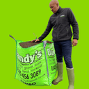 A person in a black jacket and green boots stands beside a large bag of Dandy's Topsoil & Landscape Supplies Compost Peat Free Multi Purpose Welsh, resting one hand on the bag, set against a bright green background.