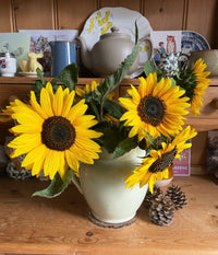 The Sunflower Waooh! by Bishy Barnabees Cottage Garden Ltd features bright yellow sunflowers in a cream-colored jug on a wooden table, with teapots, mugs, plates, greeting cards, and two pinecones as accents.