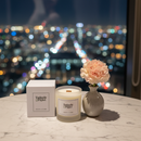 A Tadpole & Piglet Earl Grey & Cucumber Scented Candle and its box rest on a marble table beside a vase with a pink carnation, with blurred colorful city lights in the background.