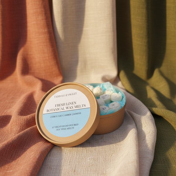 A round Tadpole & Piglet Fresh Linen Botanical Wax Melts box sits open on beige, rust, and olive fabric, revealing white floral wax melts. The label lists citrus, lily, amber, and jasmine scents.
