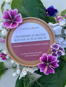 A round container of Tadpole & Piglet Cranberry & Orange Botanical Wax Melts is surrounded by fresh leaves and colorful flowers. The label notes cranberry, orange, cinnamon, and jasmine for a festive home fragrance.