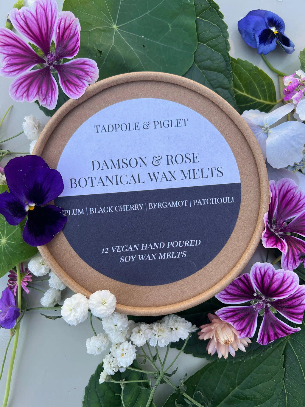 A round box of Tadpole & Piglet’s Damson & Rose Botanical Wax Melts, surrounded by colorful flowers and green leaves, features a fruity floral scent with plum, black cherry, bergamot, and patchouli notes. Contains 12 vegan botanical melts.