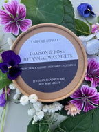 A round box of Tadpole & Piglet’s Damson & Rose Botanical Wax Melts, surrounded by colorful flowers and green leaves, features a fruity floral scent with plum, black cherry, bergamot, and patchouli notes. Contains 12 vegan botanical melts.
