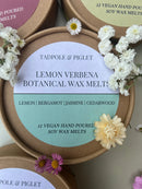 A round Tadpole & Piglet Lemon Verbena Botanical Wax Melts tin is surrounded by small white and yellow flowers. The label features a citrus aroma with lemon, bergamot, jasmine, and cedarwood notes.