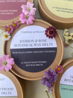 A round Tadpole & Piglet Damson & Rose Botanical Wax Melts tin is shown among other tins and colorful flowers. The label suggests a fruity floral scent with notes of plum, black cherry, bergamot, and patchouli.