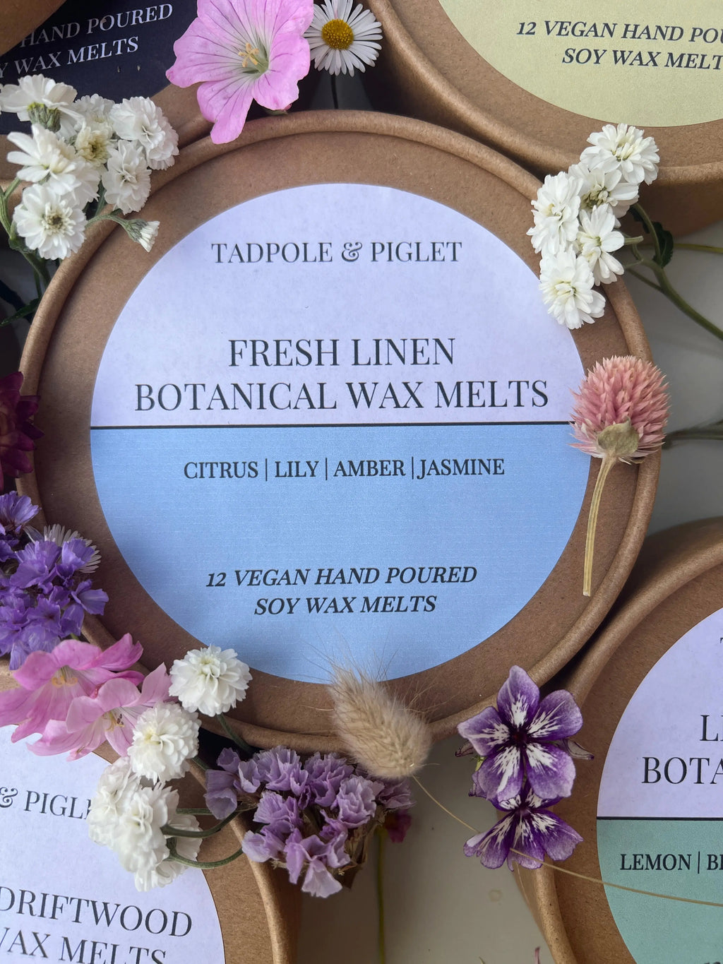 A round Tadpole & Piglet Fresh Linen Botanical Wax Melts container sits among colorful flowers, its label highlighting powdery floral scents of citrus, lily, amber, jasmine, with 12 soy wax melts inside.