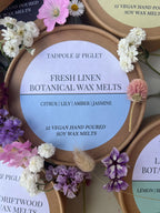 A round Tadpole & Piglet Fresh Linen Botanical Wax Melts container sits among colorful flowers, its label highlighting powdery floral scents of citrus, lily, amber, jasmine, with 12 soy wax melts inside.