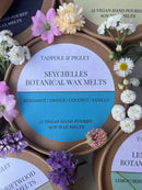 A Tadpole & Piglet Seychelles Botanical Wax Melts container sits amid colorful flowers. Its label highlights coconut and vanilla scents. Other Tadpole & Piglet botanical wax melts containers appear in the background.