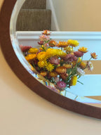 A round mirror reflects a bouquet of Bishy Barnabees Cottage Garden’s Strawflower Helichrysum Swiss Giant Mix, featuring yellow, orange, and pink blooms, placed by a white staircase with carpeted steps in the background.