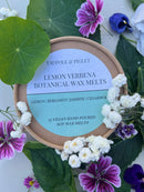 A round Tadpole & Piglet Lemon Verbena Botanical Wax Melts container sits on green leaves, surrounded by purple and white flowers. The label features a fresh citrus scent with lemon, bergamot, jasmine, and cedarwood notes; these melts are vegan.