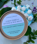 A round container of Tadpole & Piglet Lemon Verbena Botanical Wax Melts is displayed with white wax melts, green leaves, and purple flowers, evoking the fresh citrus aroma of lemon, bergamot, jasmine, and cedarwood.