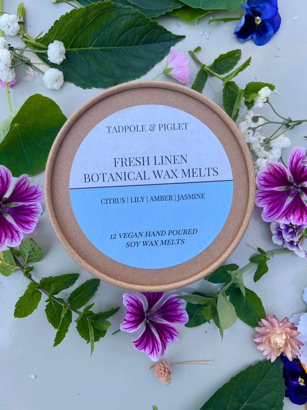 A round Tadpole & Piglet Fresh Linen Botanical Wax Melts container sits on a leafy, floral surface. Its label features powdery floral scents: citrus, lily, amber, and jasmine.