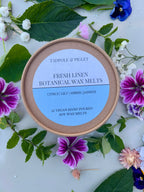 A round Tadpole & Piglet Fresh Linen Botanical Wax Melts container sits on a leafy, floral surface. Its label features powdery floral scents: citrus, lily, amber, and jasmine.