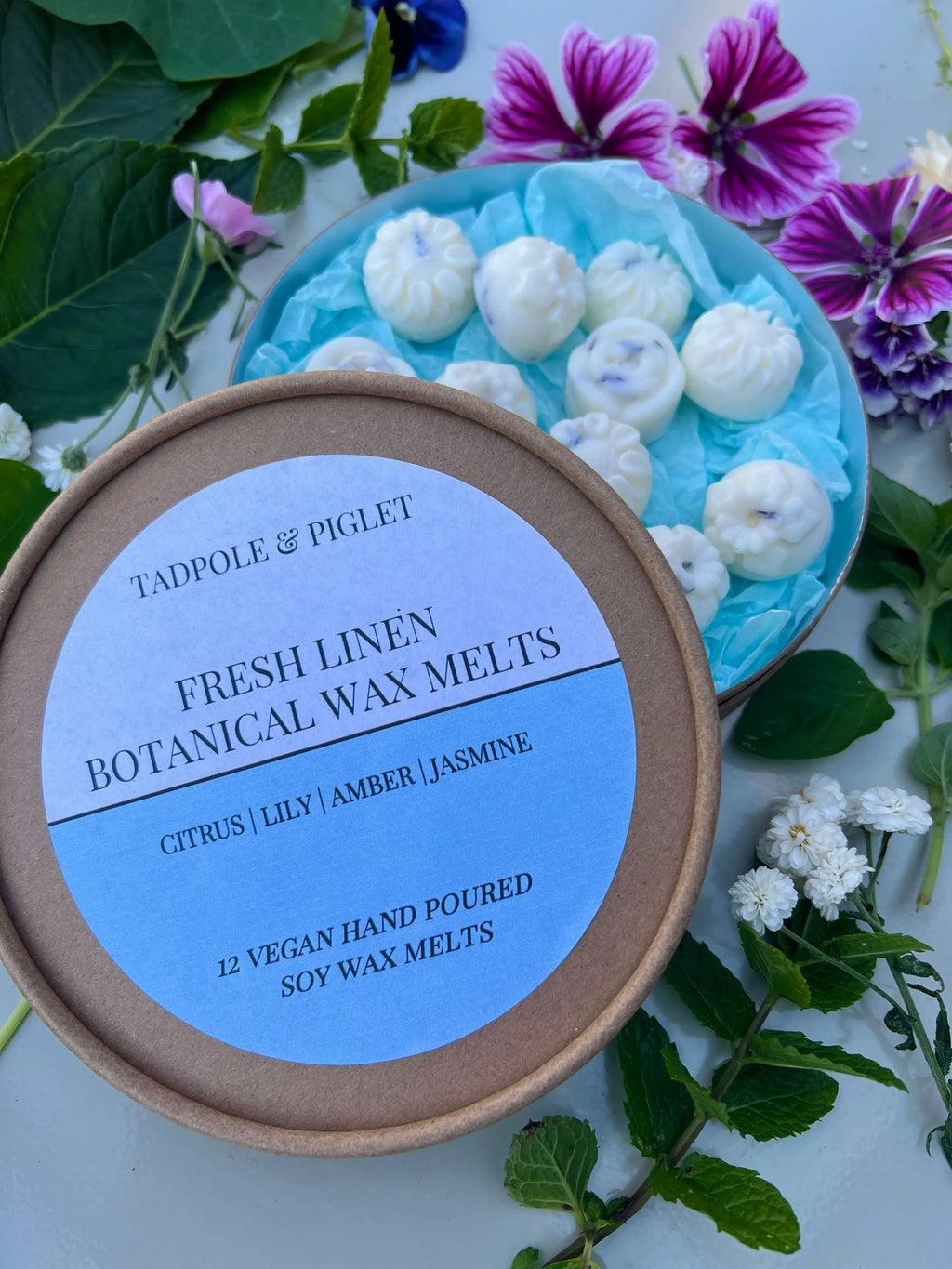 A round Tadpole & Piglet Fresh Linen Botanical Wax Melts container rests on green leaves and flowers, suggesting a powdery floral scent. Beside it, a dish displays white, flower-shaped botanical wax melts.