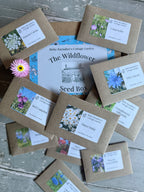 A variety of brown seed packets labeled with wildflower names surrounds The Wildflower Seed Box by Bishy Barnabees Cottage Garden Ltd, topped with a pink daisy on rustic wood—a charming addition to any British garden.