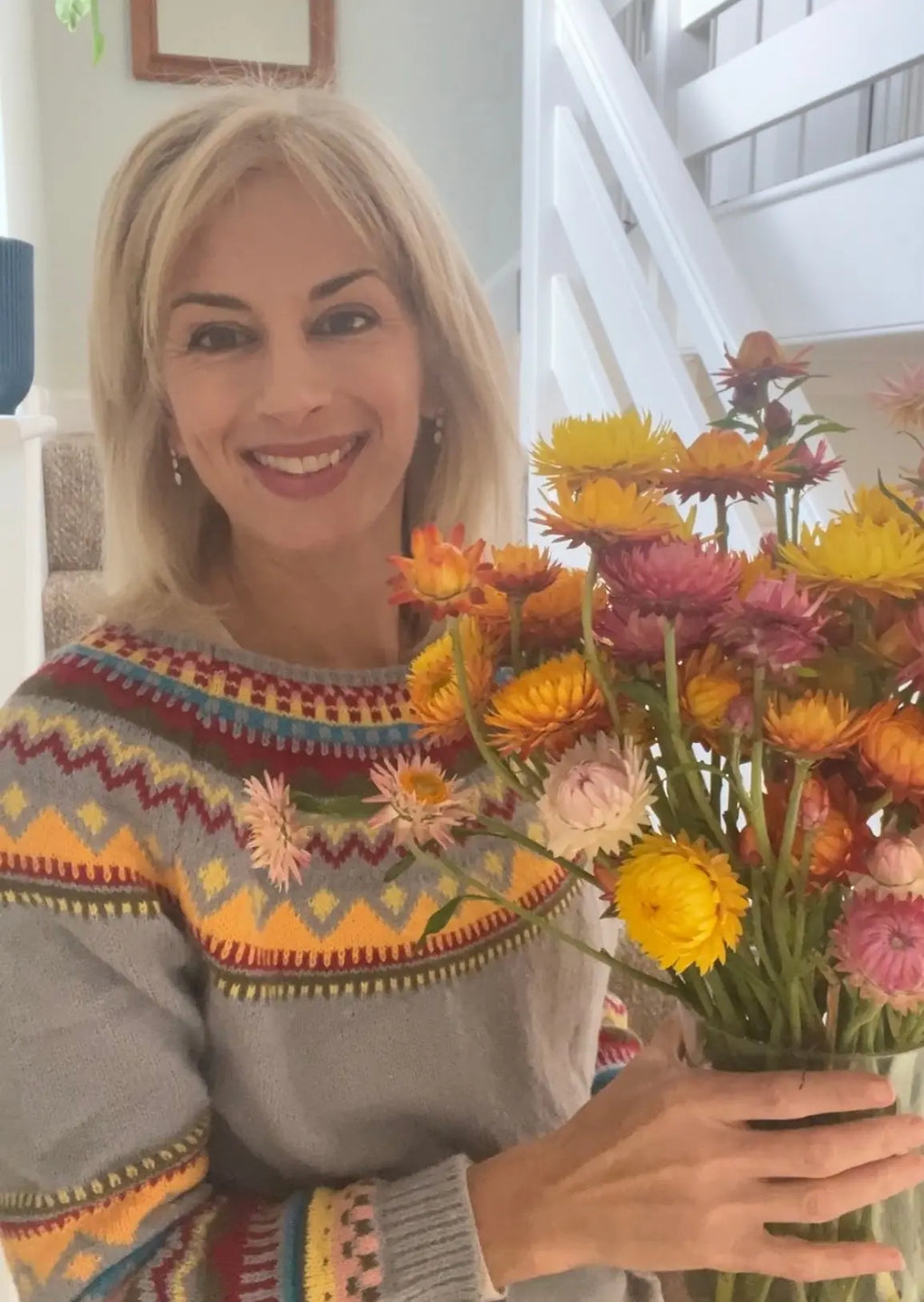 A smiling blonde woman in a colorful sweater holds a vibrant bouquet of Bishy Barnabees Cottage Garden Strawflower Helichrysum Swiss Giant Mix near a white staircase indoors.