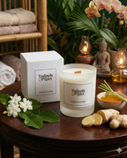 A Tadpole & Piglet Lemongrass & Ginger Scented Candle sits on a wooden table with its white box, surrounded by lemongrass, ginger, and white flowers. Greenery, towels, a Buddha statue, and the lit natural soy wax candle are in the background.