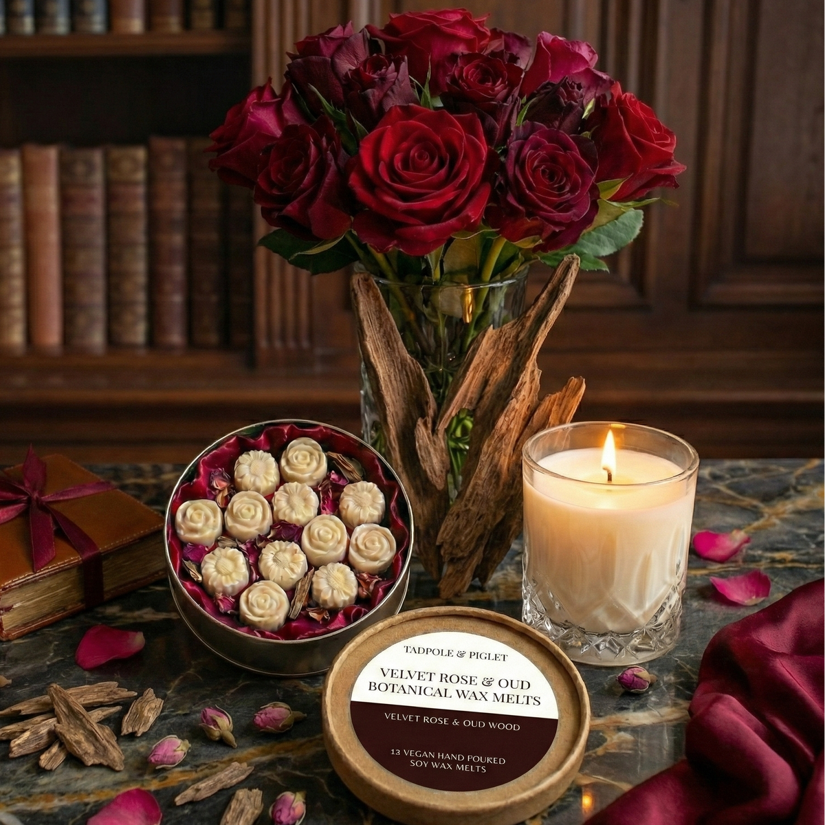 A marble table displays a lit candle, Tadpole & Piglet Velvet Rose & Oud Botanical Wax Melts tin, a vase of red roses, a gift-wrapped book, rose petals, and wood pieces with a bookshelf in the background.