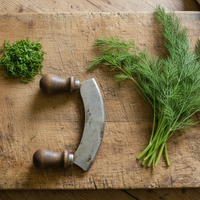 A mezzaluna knife rests on a wooden board with chopped herbs—great for pickling—and fresh dill sprigs, perfect for planting Dill Bouquet seeds from Bishy Barnabees Cottage Garden, beside a beige cloth.