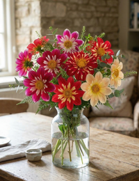 A clear vase holds a vibrant Dahlia Yankee Doodle Dandy Mix bouquet from Bishy Barnabees Cottage Garden, with lush greenery, on a wooden table by stone walls and a window—perfect bee-friendly flowers.