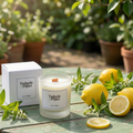 A Tadpole & Piglet Lemon Verbena Scented Candle sits on a green outdoor table with its box, lemons, lemon slices, leaves, potted plants, and sunlight in the background.