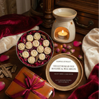On a wooden table, Tadpole & Piglet’s Velvet Rose & Oud Botanical Wax Melts—rose-shaped and hand-poured—are displayed with a lit wax warmer, petals, and a wrapped gift tied with red ribbon.