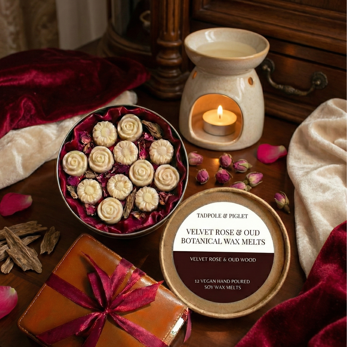 On a wooden table, Tadpole & Piglet’s Velvet Rose & Oud Botanical Wax Melts—rose-shaped and hand-poured—are displayed with a lit wax warmer, petals, and a wrapped gift tied with red ribbon.