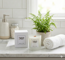 A Lime, Basil & Mandarin Scented Candle by Tadpole & Piglet sits in a glass jar on a bathroom counter beside its box, a potted fern, a rolled white towel, and a soap dispenser against a tiled wall.