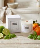 A Tadpole & Piglet Lime, Basil & Mandarin Scented Candle with a wooden wick sits by its white box, surrounded by fresh limes, basil leaves, and mandarins on a light surface under soft natural light.