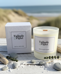 A Tadpole & Piglet Rock Salt & Driftwood Scented Candle with a wooden wick sits by its box on a white surface, surrounded by sea salt, driftwood, stones, and lavender, evoking sand dunes and ocean for a coastal-inspired aroma.