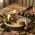 A cozy table features Tadpole & Piglet’s Wild Fig & Cassis Botanical Wax Melts shaped like flowers, a lit candle in a ceramic holder, figs, pinecones, leafy branches, and a round wooden tray on a brown cloth with warm sunlight streaming in.
