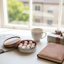 A round tin of white chocolates, a mug, a wrapped gift, Tadpole & Piglet’s Havana Nights Botanical Wax Melts, a newspaper, and a floral notebook are arranged on a white desk by a sunlit window with trees and buildings outside.