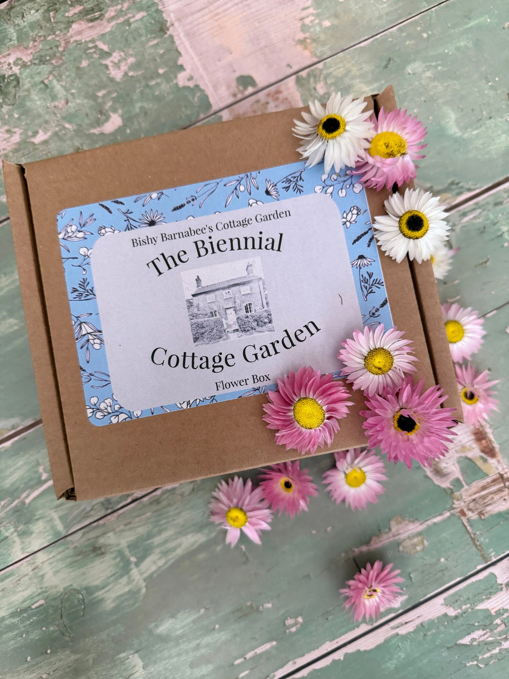 A brown box labeled The Biennial Cottage Garden Flower Box by Bishy Barnabees Cottage Garden rests on a rustic green surface, adorned with pink and white daisy-like flowers—ideal for heritage biennials or premium biennial flower seeds.