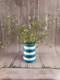 A blue and white striped vase displays Bishy Barnabees Cottage Garden Ltd's Gypsophila Pink, with delicate green stems and tiny white flowers, set against a rustic, weathered pink wooden background.