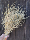 A hand holds a bundle of Lepidium sativum Dried by Bishy Barnabees Cottage Garden Ltd with light-colored wildflowers, set against a dark wooden background.