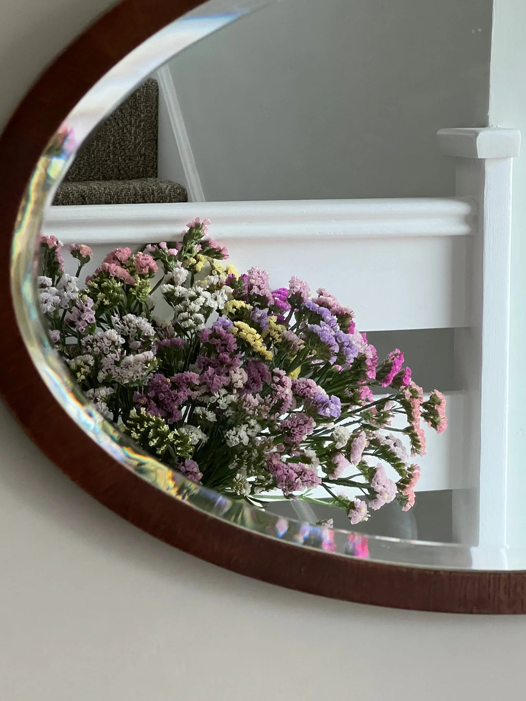 A round mirror on a white wall near a staircase reflects a bouquet of colorful Statice Hipster Mixed blooms by Bishy Barnabees Cottage Garden in shades of pink, purple, yellow, and white.