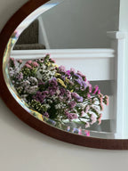 A round mirror on a white wall near a staircase reflects a bouquet of colorful Statice Hipster Mixed blooms by Bishy Barnabees Cottage Garden in shades of pink, purple, yellow, and white.