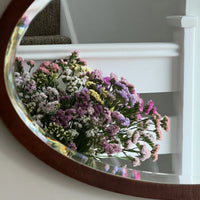 A round mirror on a white wall near a staircase reflects a bouquet of colorful Statice Hipster Mixed blooms by Bishy Barnabees Cottage Garden in shades of pink, purple, yellow, and white.