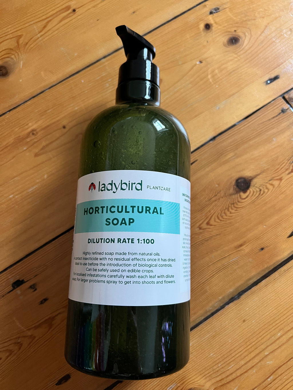 A green pump bottle labeled ladybirdplantcare.co.uk Horticultural Soap sits on a wooden floor. The label notes a 1:100 dilution rate and describes it as a contact insecticide from natural oils for plant care.