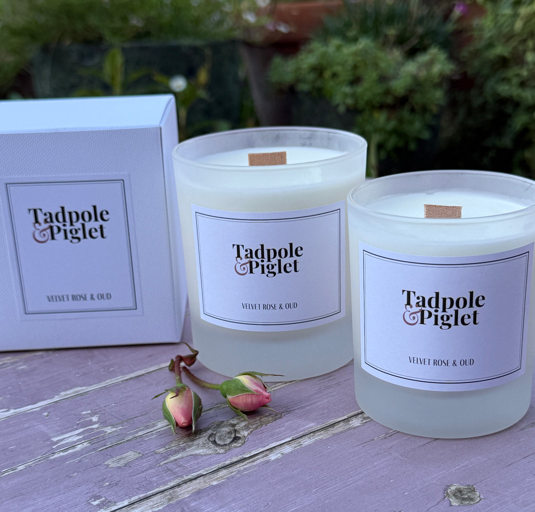 Two Tadpole Piglet candles on a wooden surface with a box and flowers in the background.