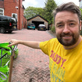 A man in a yellow DADDY T-shirt, smiling and holding a garden tool in the driveway, stands next to a green bag of Dandy's Topsoil & Landscape Supplies' Topsoil Welsh VegeGrow® for vegetables; cars and a red brick house are in the background.