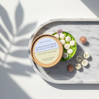 A round Tadpole & Piglet box labeled “Lemongrass & Ginger Botanical Wax Melts” sits open on a marble tray, revealing flower-shaped melts. A small flower and sunlight evoke a luxurious, fresh Thai spa atmosphere.