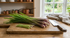 A bunch of fresh lemongrass stalks on a rustic wooden chopping board, next to a large knife, with a kitchen tea towel and copper pots in the background by a window.