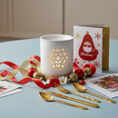 A festive table features gold cutlery, red and gold ornaments, ribbon, a Tadpole & Piglet White Mandala Cut Out Wax Burner as a candle holder, and Christmas cards, including one illustrated with Santa.
