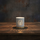 The Tadpole & Piglet White Mandala Cut Out Wax Burner, with intricate patterns, sits on a wooden table casting warm light onto the surface. The background is dark and textured.