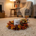 A white Tadpole & Piglet Myrrh & Tonka Scented Candle rests on a beige carpet with autumn leaves and berries, set against a background of a cozy armchair, stool, bookshelf, and soft lighting for luxurious home fragrance.