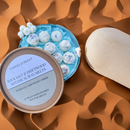 A round Tadpole & Piglet Rock Salt & Driftwood Botanical Wax Melts box sits open on an orange patterned surface, showing floral-adorned wax melts inside. A beige oval stone is placed next to the box.