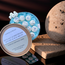 An open tin of Tadpole & Piglet Rock Salt & Driftwood Botanical Wax Melts displays decorative white, hand-poured wax melts with blue accents, accompanied by a textured stone sphere and a rectangular stone slab.