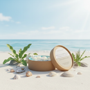 A round, brown box of Tadpole & Piglet’s Rock Salt & Driftwood Botanical Wax Melts rests open on a sandy beach with shells and flowers. Enjoy its serene scent solo or pair it with other botanical candles for relaxing vibes anywhere.